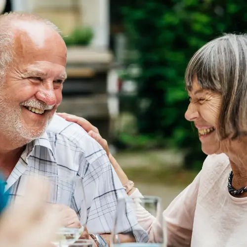 Deux personnes âgées sourient et rient ensemble lors d'un repas en extérieur. L'homme avec une barbe grise porte une chemise à rayures, tandis que la femme a les cheveux courts et argentés. Ils semblent partager un moment joyeux et complice, entourés de verdure.