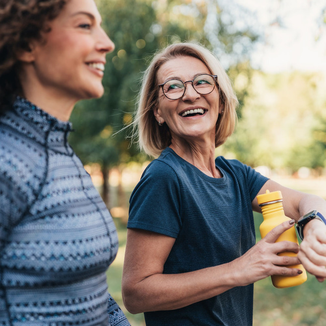 Deux femmes souriantes marchent ensemble dans un parc, entourées d'arbres. L'une porte un haut à motifs et l'autre une t-shirt simple avec des lunettes. La femme plus âgée tient une bouteille d'eau jaune. Elles semblent heureuses et en bonne humeur.