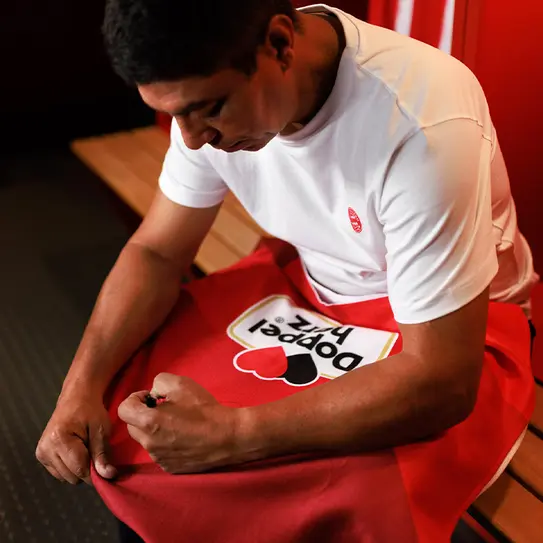 Un homme en t-shirt blanc est assis sur un banc dans un vestiaire, en train de réparer un vêtement rouge avec un logo en forme de cœur. Il semble concentré sur sa tâche. La lumière dans la pièce est douce et ambiante.