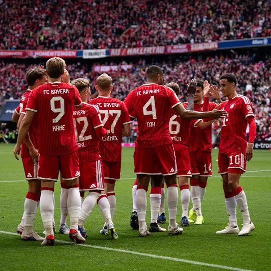 Des joueurs du FC Bayern Munich se regroupent, célébrant un moment sur le terrain. Ils portent des maillots rouges et blancs, avec des numéros affichés. En arrière-plan, une foule animée soutient l'équipe dans un stade.