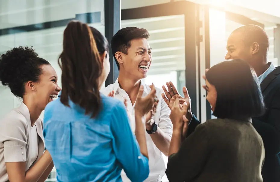 Un groupe de six personnes souriantes se rassemble dans un espace de travail. Elles échangent des rires et des applaudissements, créant une atmosphère joyeuse et amicale. La lumière du jour éclaire la scène, soulignant leur convivialité.