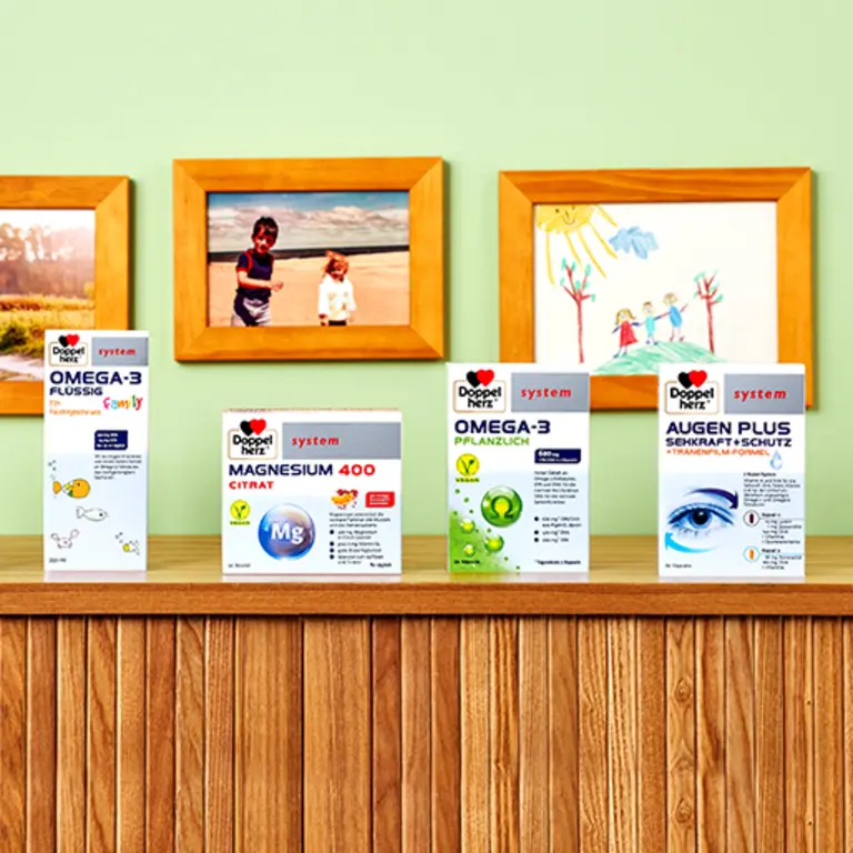 Quatre boîtes de produits de santé sont alignées sur une étagère en bois. Les boîtes sont étiquetées "OMEGA-3 Flüssig", "MAGNESIUM 400 CITRAT", "OMEGA-3 Pflanzlich" et "AUGEN PLUS Sehkraft-Schutz". En arrière-plan, des dessins d'enfants et des photos en couleur sont accrochés au mur.