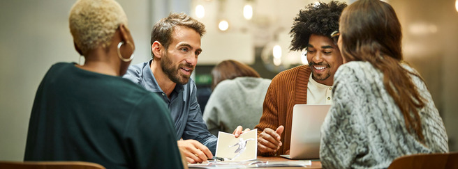 Un groupe de quatre personnes est assis autour d'une table. Ils discutent et échangent des idées, avec un ordinateur portable et des documents devant eux. L'atmosphère est collaborative et informelle.