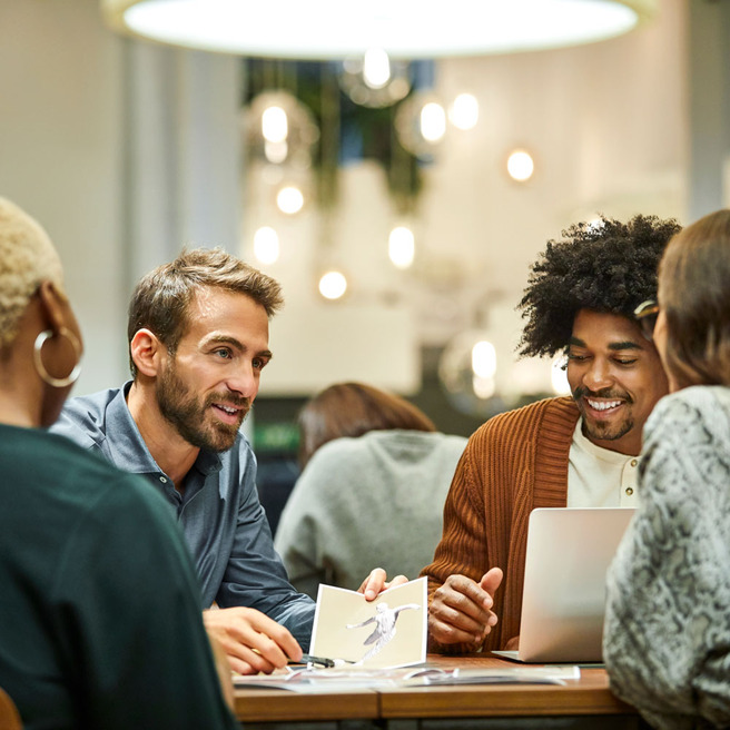 Un groupe de personnes discute autour d'une table, avec un homme souriant portant une chemise décontractée. Ils semblent engagés dans une conversation collaborative, avec des documents et un ordinateur portable sur la table. L'environnement est moderne et lumineux.