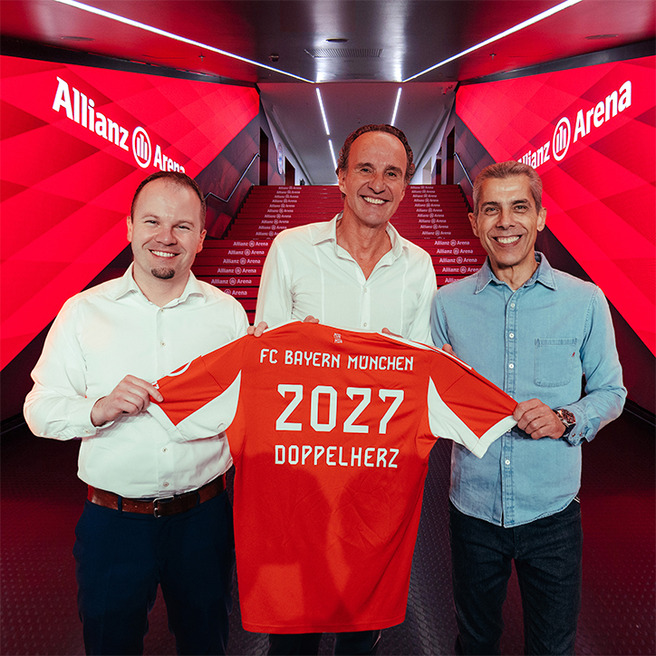 Trois hommes souriants se tiennent sous l’enseigne de l’Allianz Arena, tenant une maillot de football rouge du FC Bayern Munich. Le maillot affiche "2027" et "DOPPELHERZ" au dos. L'atmosphère est festive, suggérant un événement important pour le club.