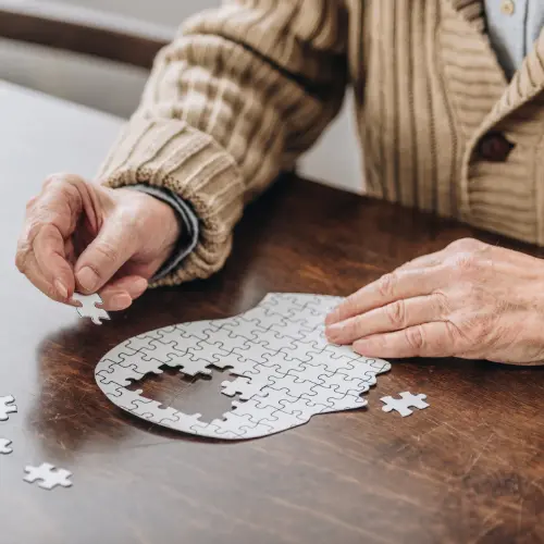 Une personne âgée assemble un puzzle sur une table en bois. Elle tient une pièce de puzzle dans une main et cherche à l'ajouter à un motif en cours. Des pièces de puzzle sont éparpillées autour.