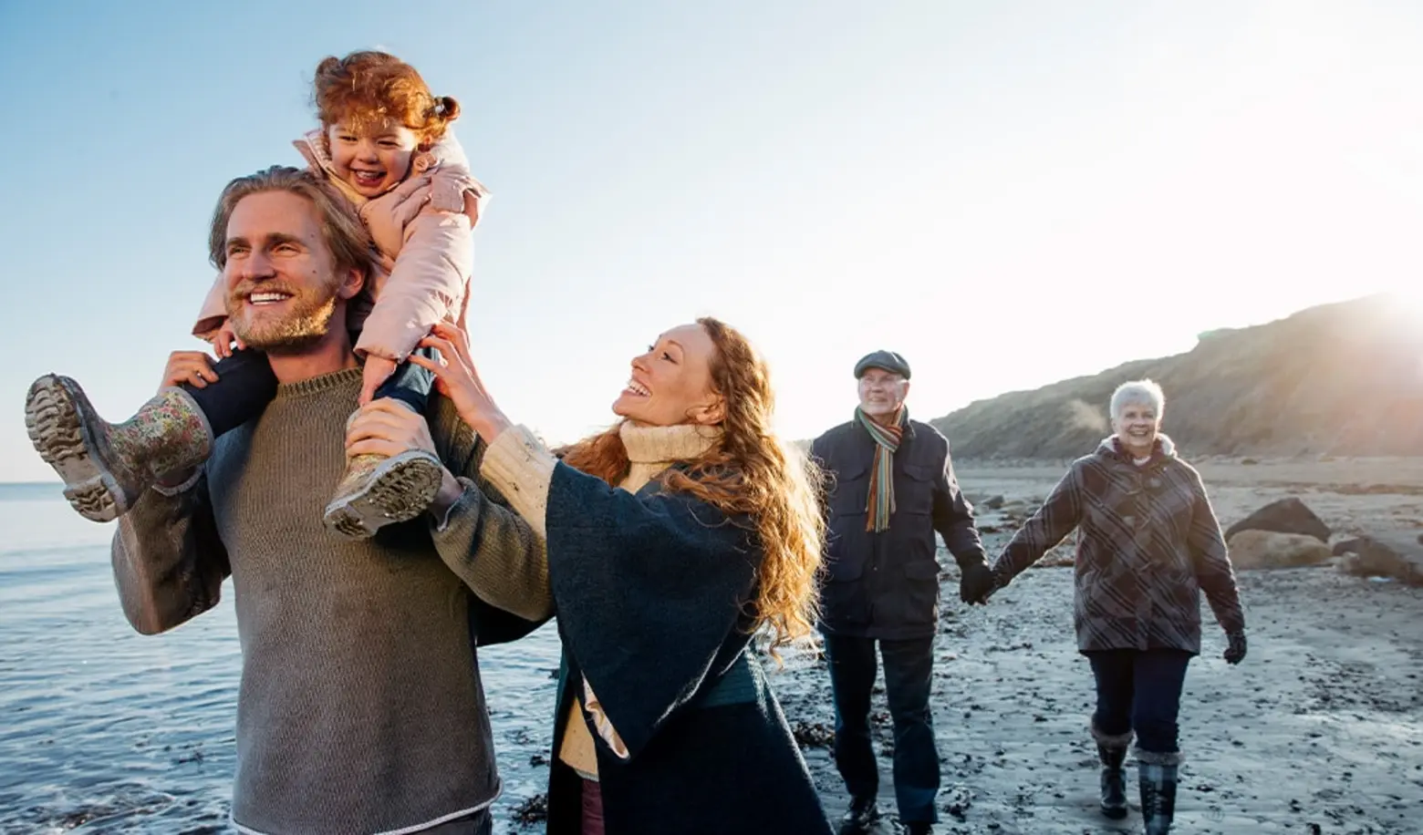 Une famille souriante se promène sur la plage. Un homme porte une petite fille sur ses épaules, tandis qu'une femme marche à ses côtés. À l'arrière-plan, un couple plus âgé se tient la main. Le soleil se lève sur l'horizon, créant une ambiance chaleureuse.