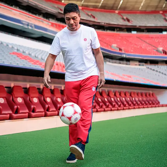 Un homme souriant, portant un t-shirt blanc et un pantalon rouge, jongle avec un ballon de football sur un terrain dans un stade vide. Les gradins en arrière-plan sont décorés de sièges rouges.