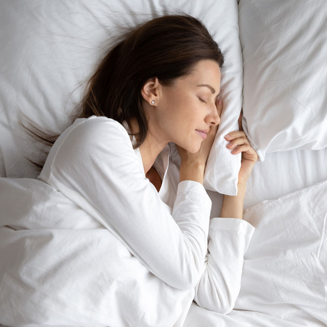 Une femme est allongée sur un lit, apparaissant confortable et au repos. Elle porte un haut blanc et a les cheveux bruns, légèrement ondulés. Son visage exprime la sérénité tandis qu'elle se tourne vers un côté sur les oreillers.