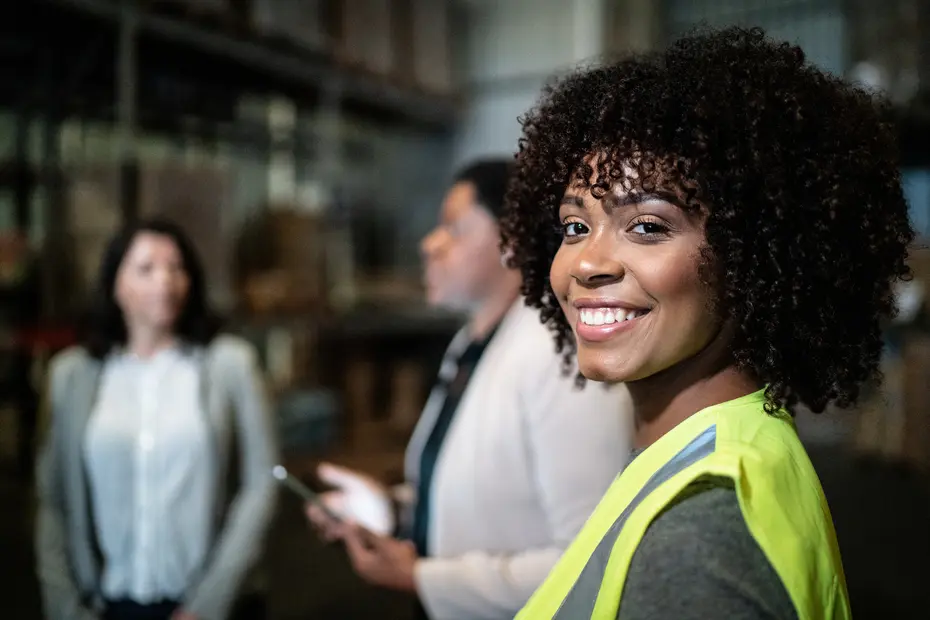 Une femme souriante portant un gilet de sécurité se tient en avant-plan dans un entrepôt. Deux autres femmes, floues en arrière-plan, discutent. L'environnement semble professionnel et axé sur le travail.