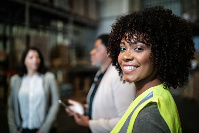Une femme souriante portant un gilet de sécurité se tient en avant-plan dans un entrepôt. Deux autres femmes, floues en arrière-plan, discutent. L'environnement semble professionnel et axé sur le travail.