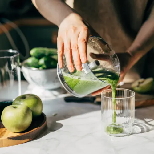 Une main verse un smoothie vert dans un verre transparent. Sur la table, des pommes vertes et des concombres sont visibles, ainsi qu'un mixeur et un bol. La lumière naturelle met en valeur la fraîcheur des ingrédients.