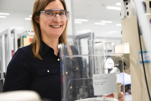 Une femme souriante portant des lunettes de protection se tient près d'un équipement de laboratoire. Elle porte une chemise noire et se trouve dans un environnement de recherche scientifique.
