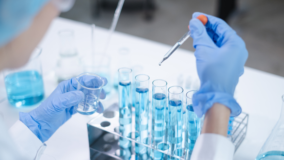 Un scientifique en blouse blanche et gants en latex tient un bécher dans une main et une pipette dans l'autre, ajoutant un liquide bleu dans des éprouvettes disposées sur une étagère. Le laboratoire est bien éclairé, avec des contenants de liquides visibles en arrière-plan.