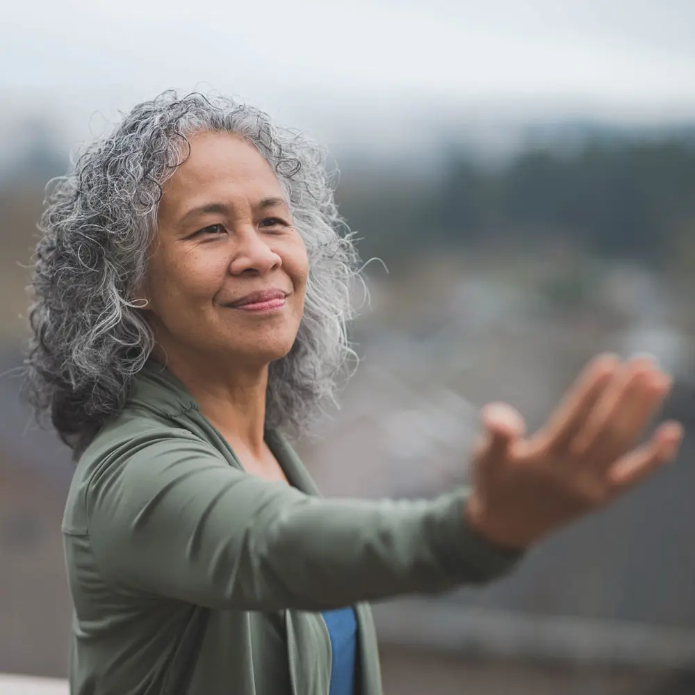 Une femme d'âge moyen avec des cheveux bouclés et gris se tient debout, le bras légèrement tendu vers l'avant. Elle affiche un sourire serein, dans un environnement extérieur légèrement brumeux.
