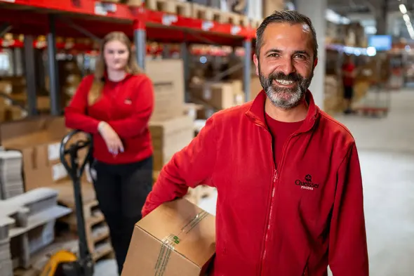 Deux employés de magasin dans un entrepôt. Un homme souriant tient une boîte, tandis qu'une femme en arrière-plan se tient près d'un transpalette. Tous deux portent des vêtements rouges, avec des rangées de caisses en arrière-plan.