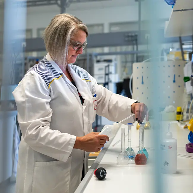 Une scientifique en blouse de laboratoire utilise une pipette pour manipuler des liquides dans des flacons, entourée d'équipements de laboratoire.
