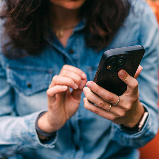 Une personne portant une chemise en denim regarde son téléphone avec une main, tout en touchant l'écran avec l'autre main. Les cheveux sont lâchés et la concentration est visible sur son visage.