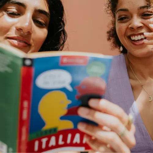 Deux femmes regardent un livre sur l'italien. L'une lit à voix haute, tandis que l'autre sourit et semble engagée. Le livre est coloré, avec des illustrations de silhouettes. L'arrière-plan est de couleur chaude.