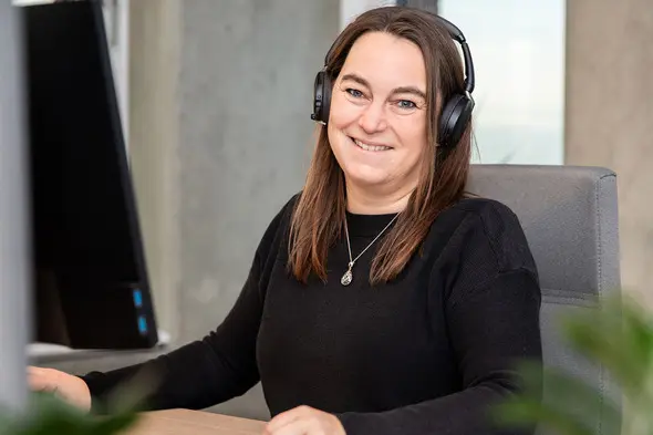 Femme souriante portant un casque audio, assise à un bureau devant un ordinateur. Elle a les cheveux longs et bruns, et porte un pull noir. En arrière-plan, on aperçoit des murs en béton et des éléments de bureau.