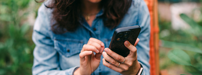 Une personne avec des cheveux bouclés utilise un smartphone, assise dans un environnement verdoyant. Elle porte une chemise en jean et semble absorbée par l'écran de son appareil.