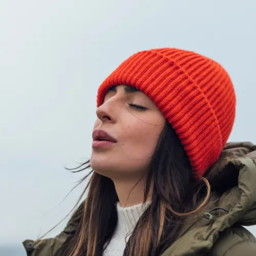 Une femme portant un bonnet orange se tient à l'extérieur, les yeux fermés, semblant profiter d'un moment de calme. Elle est vêtue d'une veste épaisse et d'un pull clair, avec un arrière-plan flou évoquant un environnement naturel.