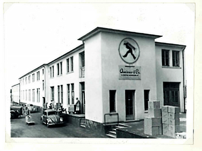Bâtiment d'une usine en noir et blanc, avec un logo représentant un homme en mouvement sur la façade. Des personnes sont visibles à l'extérieur, et des caisses sont empilées près d'une voiture ancienne.