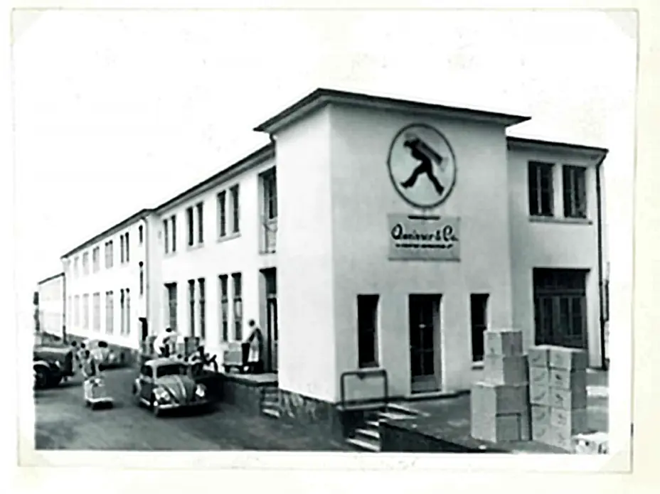 Bâtiment d'une usine en noir et blanc, avec un logo représentant un homme en mouvement sur la façade. Des personnes sont visibles à l'extérieur, et des caisses sont empilées près d'une voiture ancienne.