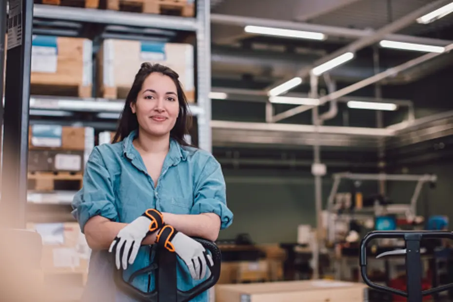 Une femme souriante en tenue de travail se tient devant un chariot élévateur dans un environnement d'entrepôt. Elle porte des gants de sécurité et une chemise en jean, montrant une attitude confiante au milieu de caisses et d'équipements.