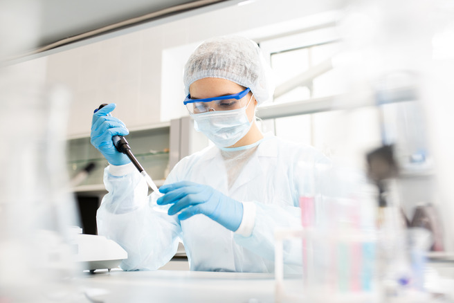 Une scientifique en blouse blanche, portant des gants et un masque, utilise un micropipette dans un laboratoire. Elle est concentrée sur son travail, entourée d'équipements de laboratoire.