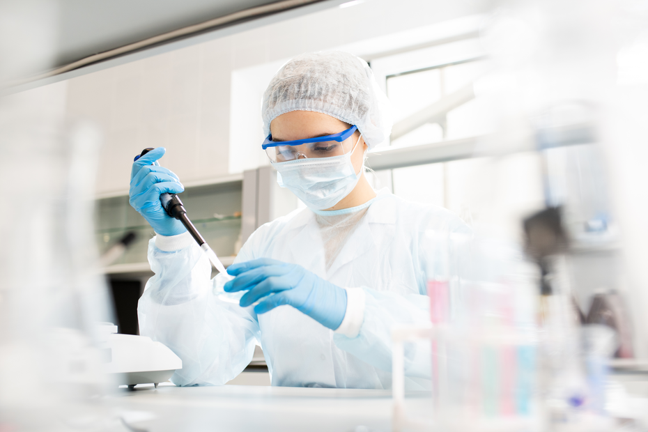 Une scientifique en blouse blanche, portant des gants et un masque, utilise un micropipette dans un laboratoire. Elle est concentrée sur son travail, entourée d'équipements de laboratoire.
