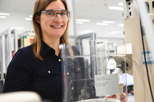 Une femme souriante portant des lunettes de protection se tient près d'un équipement de laboratoire. Elle porte une chemise noire et se trouve dans un environnement de recherche scientifique.