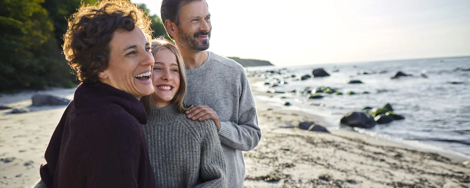 Trois personnes souriantes se tenant sur une plage. Deux femmes, l'une avec des cheveux bouclés, l'autre avec des cheveux longs, portent des pulls. Un homme les entoure, affichant un sourire. En arrière-plan, la mer et des rochers sont visibles sous un ciel ensoleillé.